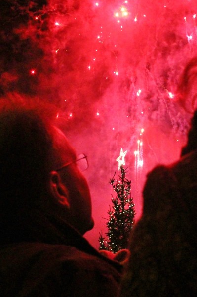 A man looks up at the night's sky, illuminated by red fireworks.